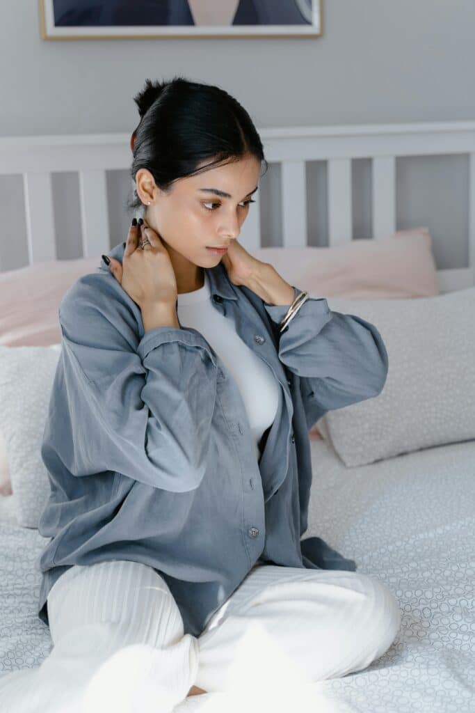Young woman sitting on bed massaging her neck for self-care and relaxation.